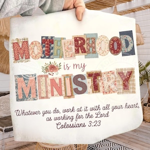 May include: A white decorative pillow with the words "MOTHERHOOD is my MINISTRY" in colorful, patchwork-style letters. The pillow also features a floral design and a Bible verse, Colossians 3:23.