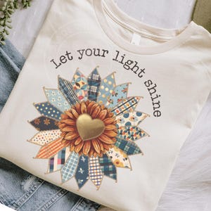 May include: Cream-colored t-shirt featuring a sunflower design with a gold heart center and patchwork petals in shades of blue, orange, and brown. The text "Let your light shine" is printed above the sunflower in black. The shirt is made of soft material.