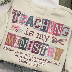 May include: Cream-colored sweatshirt with the words "TEACHING is my MINISTRY" in colorful, patchwork-style letters. A pencil and bow graphic is included. The text "Whatever you do, work at it with all your heart, as working for the Lord Colossians 3:23" is also present.