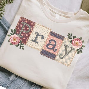 Może przedstawiać: Kremowa koszulka z napisem "PRAY" w patchworkowym wzorze. Każda litera znajduje się na innym kwadracie z wzorem, z kwiatowymi akcentami i obszytą krawędzią. Koszulka ma klasyczny okrągły dekolt.