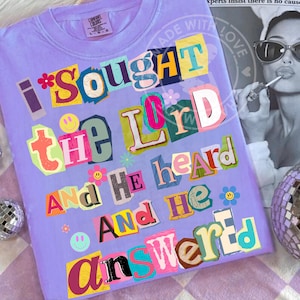 Puede incluir: Una camiseta lila Comfort Colors con el texto "I sought the Lord and He heard and He answered" en un diseño de letras coloridas recortadas. La camiseta presenta caras sonrientes y gráficos de flores.