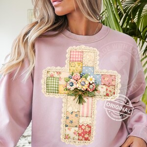 May include: A light purple sweatshirt featuring a cross design. The cross is made of patchwork fabric in various floral and gingham patterns, trimmed with lace, and adorned with a bouquet of flowers. The word "ORIGINAL" is stamped on the lower right.