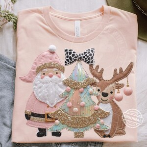 May include: A light pink t-shirt featuring a festive Christmas design. The design includes a Santa Claus, a Christmas tree, and a reindeer, all adorned with glitter and pastel colors. A black and white bow is above the tree.