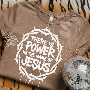 May include: A brown t-shirt featuring the white text "THERE IS POWER IN THE NAME OF JESUS" within a white crown of thorns. The shirt has a crew neck and short sleeves. A disco ball is visible in the bottom right corner.