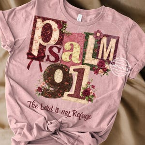 May include: A light pink t-shirt featuring the text "Psalm 91" in a patchwork design with floral accents. The words "The Lord is my Refuge" are printed below the numbers. The shirt has short sleeves and a crew neck.