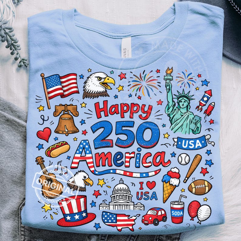 May include: Light blue t-shirt with a colorful graphic celebrating the 250th anniversary of America. The design includes the American flag, Statue of Liberty, fireworks, and the text "Happy 250 America".