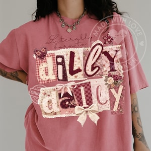 May include: Dusty rose t-shirt with the text "Literally born to dilly dally" in a patchwork design. The design includes floral patterns, bows, and a heart. The shirt has short sleeves and a crew neck.