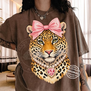 Puede incluir: Camiseta marrón con una ilustración de leopardo con un lazo rosa, collar de perlas y un colgante en forma de corazón rosa. El leopardo tiene ojos verdes y un pelaje manchado. La camiseta tiene las palabras "HAND MADE" y "ORIGINAL".