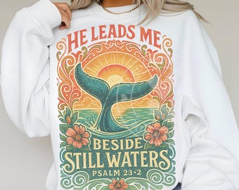 May include: White sweatshirt with a colorful graphic featuring a whale tail, sun, and floral elements. The text reads "HE LEADS ME BESIDE STILL WATERS PSALM 23:2" in a decorative font. The design incorporates shades of teal, orange, and green.