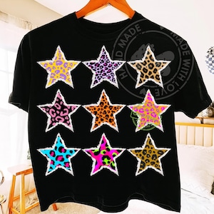 Könnte beinhalten: Schwarzes T-Shirt mit neun sternförmigen Designs mit Tiermustern in verschiedenen Farben, darunter Gelb, Lila, Braun, Pink, Orange, Blau und Grün. Jeder Stern hat einen weißen, zerrissenen Papierrand. Das Shirt hängt an einem Holzkleiderbügel.