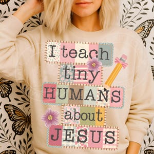 Op de afbeelding: Beige sweatshirt met de tekst "I teach tiny HUMANS about JESUS" in een patchwork-ontwerp. Voorzien van een gele potlood met een roze gum en strik, en twee paarse bloemen. Een comfortabele top.