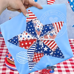 May include: A light blue pillow with a large star design. The star is made of patches with red, white, and blue patterns, including stars, stripes, and American flags. The pillow has the words "MADE WITH LOVE" and "ORIGINAL" printed on it.