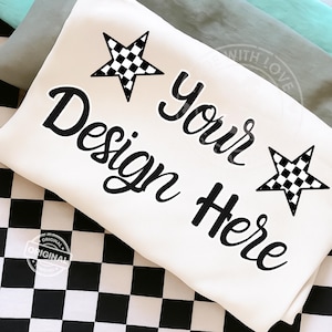 Op de afbeelding: Crèmekleurig kussen met de tekst "Your Design Here" in zwart script. Twee zwart-wit geblokte sterren staan op het kussen. Het kussen ligt op een zwart-wit geblokt oppervlak.