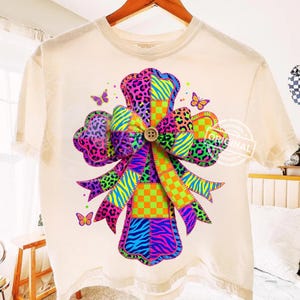 May include: A cream-coloured t-shirt with a vibrant cross design. The cross is decorated with a patchwork of animal prints, zebra stripes, and checkered patterns in pink, purple, green, and orange. Small butterfly accents add a whimsical touch.
