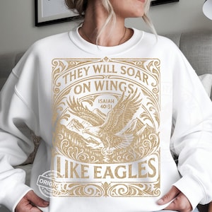 May include: White sweatshirt with a gold graphic featuring an eagle soaring over mountains. The text reads "THEY WILL SOAR ON WINGS LIKE EAGLES" with the Bible verse Isaiah 40:31.