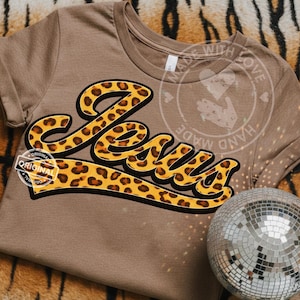 May include: A tan t-shirt with the word "Jesus" in a bold, leopard print design. The text is outlined in black and has a yellow background. The shirt has short sleeves and a round neckline.