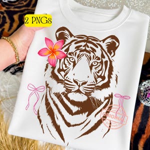 Puede incluir: Sudadera blanca con un diseño de tigre marrón, una flor rosa y naranja y lazos rosas. El texto "2 PNGs" está en un rectángulo amarillo. Un sello rosa dice "ORIGINAL".