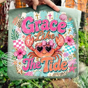May include: A green t-shirt featuring a crab wearing heart-shaped sunglasses and a pink bow. The shirt has the words "Grace Like The Tide" in pink and orange, with floral and seashell accents. The design includes crosses and a checkered pattern.