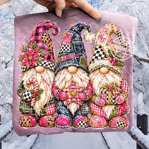 Könnte beinhalten: Ein malvenfarbenes Sweatshirt mit einem Grafikdruck von drei Gnomen. Der linke Gnom trägt eine rosa karierte Mütze mit einem Weihnachtsstern, der mittlere eine blaue Jeans-Mütze und der rechte eine florale Mütze. Sie halten eine Zuckerstange, einen Stern und ein Geschenk.