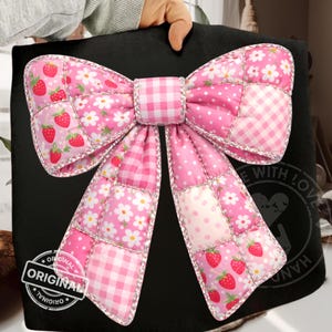 May include: A large, pink quilted bow with a patchwork design. The bow features various patterns, including strawberries, daisies, gingham, and polka dots. Silver trim outlines the bow. The word "ORIGINAL" is stamped on the bottom left.