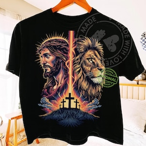 May include: Black t-shirt featuring a graphic design with two portraits: one of Jesus wearing a crown of thorns and the other of a lion. A beam of light connects the two images, with three crosses on a hill below.