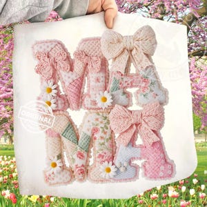 May include: A fabric sign spelling "MAMA" in pink and white patchwork letters. Each letter features a bow and daisy accents. The sign is held up against a floral background, with the word "ORIGINAL" stamped on the lower left.