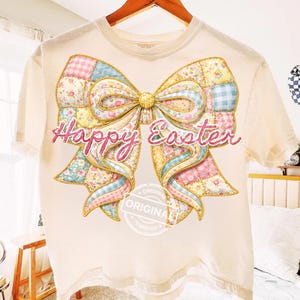 May include: A cream-coloured t-shirt with a large patchwork bow design in pastel colours, featuring floral and gingham patterns. The words "Happy Easter" are written in pink script across the bow. A circular stamp with the word "ORIGINAL" is at the bottom.