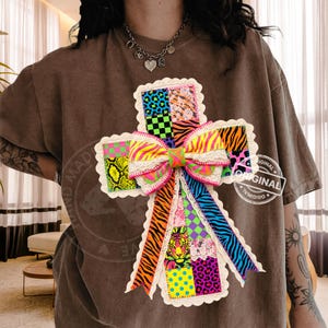 May include: A brown t-shirt with a vibrant, cross-shaped applique. The applique is a patchwork of animal prints, polka dots, and a large bow. The shirt has the words "HAND MADE" and "ORIGINAL" printed on it.