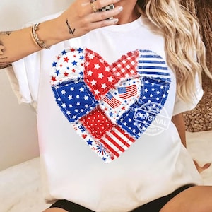 Op de afbeelding: Wit t-shirt met een patchwork hart ontwerp in rood, wit en blauw. Het hart is samengesteld uit verschillende stofpatronen, waaronder sterren, strepen en de Amerikaanse vlag. Het woord "ORIGINAL" staat op het t-shirt.