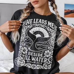 May include: Black t-shirt with a white graphic featuring a whale tail, sun, and floral elements. The text reads "HE LEADS ME BESIDE STILL WATERS PSALM 23:2". The shirt has a relaxed fit.