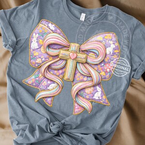 以下が含まれることがあります： 明るい青色のTシャツ。大きなカラフルなリボン模様が特徴です。リボンは金色の十字架、ピンクのハート、ウサギや卵などのイースターをテーマにした要素で飾られています。リボンは紫とピンクの配色です。