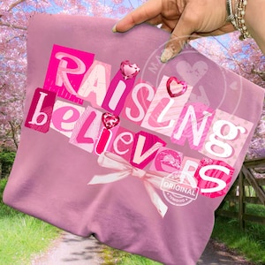 Puede incluir: Una camiseta rosa con la frase "Raising Believers" en un diseño de collage. El texto utiliza tonos rosas y blancos, con adornos en forma de corazón y un lazo blanco. La camiseta tiene un aspecto relajado e informal.