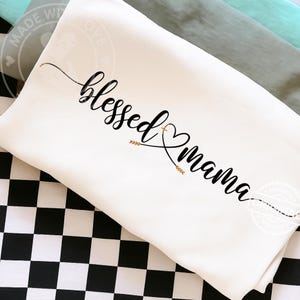 May include: A white pillow featuring the phrase "blessed mama" in black script. The design includes a heart and gold arrow accents. The pillow rests on a black and white checkered surface, with other fabric in the background.