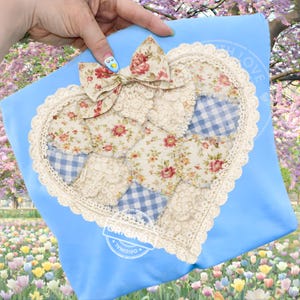 Puede incluir: Un artículo de tela azul claro con un diseño de patchwork en forma de corazón. El corazón presenta patrones florales y de cuadros, ribeteados con encaje. Un lazo floral está en la parte superior. La palabra "ORIGINAL" está estampada en la parte inferior.