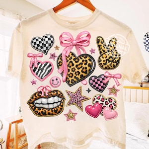 May include: Cream-colored t-shirt adorned with various patches. The patches include hearts with checkered, zebra, and leopard prints, a peace sign, lips, stars, and bows. The patches are in pink, black, and gold.