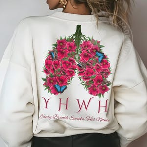 May include: Cream-colored sweatshirt featuring a floral lung design in shades of pink and green, adorned with blue butterflies. The design includes the letters YHWH and the phrase "Every Breath Speaks His Name."