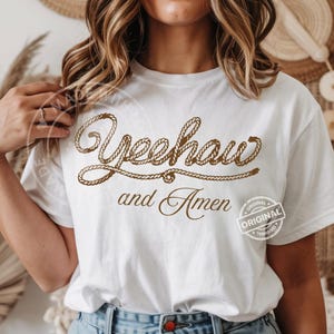 Peut inclure: T-shirt blanc avec l'inscription "Yeehaw and Amen" en marron, en forme de corde. Le t-shirt porte &eacute;galement un petit tampon "Original". Le motif est centr&eacute; sur le devant du t-shirt.