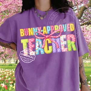 Op de afbeelding: Een paars t-shirt met de tekst "BUNNY-APPROVED TEACHER" in kleurrijke, patroonletters. Een roze strik is gecentreerd boven het woord "TEACHER". Het shirt heeft ook een "ORIGINAL" stempel.