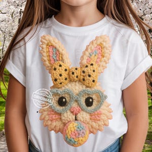 May include: White t-shirt featuring a crocheted bunny face with a bow, heart-shaped glasses, and a bubblegum bubble. The bunny has a cream-colored face, pink and blue accents, and a cookie-like bow. The word "ORIGINAL" is printed on the shirt.