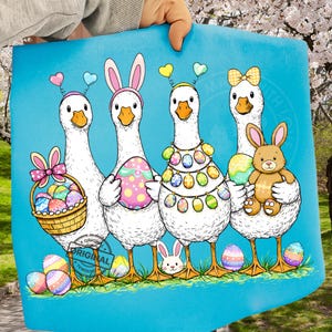 May include: A blue Easter-themed print featuring four white geese with Easter egg decorations and bunny ears. One goose holds a basket of eggs, another holds a plush bunny. The geese stand on grass with colorful eggs scattered around.