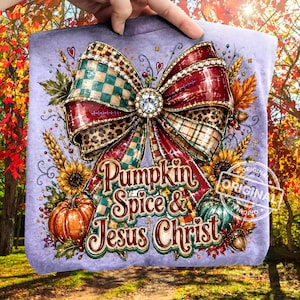 May include: A light purple t-shirt featuring a large central graphic of a decorative bow with plaid and leopard print patterns. The design includes sunflowers, wheat stalks, an orange pumpkin, and the text Pumpkin Spice & Jesus Christ.