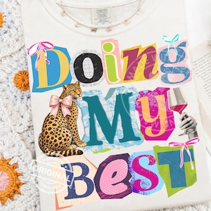 May include: Cream-colored t-shirt with the phrase "DOING MY BEST" in colorful, layered letters. A leopard with a pink bow is on the left. A hand holding a wine glass is on the right. The shirt has a Comfort Colors tag.