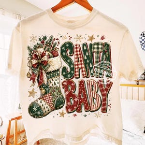 May include: A cream-colored t-shirt featuring a graphic of a Christmas stocking filled with ornaments and a large red bow. The text SANTA BABY is printed in large, patterned block letters in shades of red, green, and white, surrounded by small stars and snowflakes.