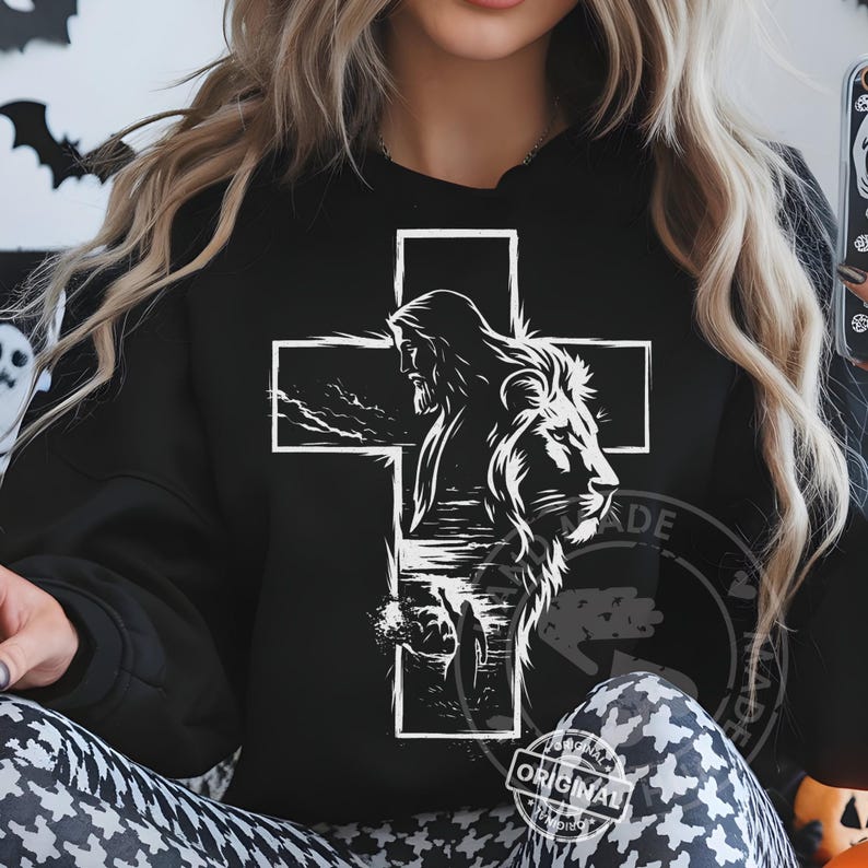 May include: Black sweatshirt featuring a white graphic of a cross with Jesus and a lion's head. The design includes a landscape scene. The word "ORIGINAL" is printed on the bottom right.