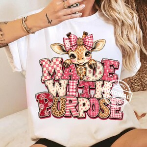 Könnte beinhalten: Ein weißes T-Shirt mit einer Grafik einer Cartoon-Giraffe, die eine rosa-weiß karierte Schleife mit einem Kreuzanhänger trägt. Der Text MADE WITH PURPOSE ist in großen, gemusterten Blockbuchstaben in Rosa-, Rot- und Beigetönen dargestellt.