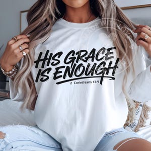 May include: White long-sleeved sweatshirt with the text "HIS GRACE IS ENOUGH" in black script. Below the text is the reference "2 Corinthians 12:9". The sweatshirt is a casual style.