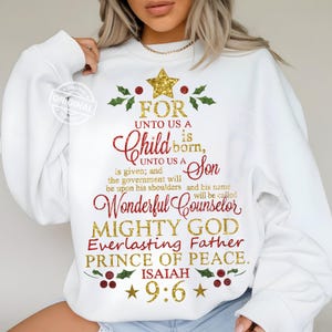 May include: White sweatshirt with a Christmas tree design made of text. The text includes the phrases "For Unto Us a Child is Born" and "Mighty God Everlasting Father Prince of Peace." The text is in red and gold glitter.