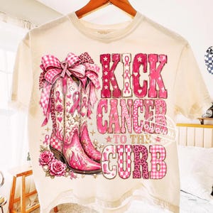 Op de afbeelding: Een crèmekleurig t-shirt met een afbeelding van twee roze cowboylaarzen versierd met een grote geruite strik en rozen. De tekst KICK CANCER TO THE CURB staat in dikke roze letters met luipaardprint en sterpatronen in het ontwerp.