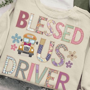 May include: Cream-colored sweatshirt with the words "BLESSED US DRIVER" in patchwork letters. A yellow school bus graphic is between the words "BLESSED" and "US". Floral and star accents are also included in the design.