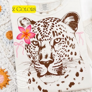 May include: Cream-colored t-shirt featuring a detailed brown leopard face with spots. A pink and orange flower adorns the leopard's head, with pink bows on either side. The text "2 COLORS" is in a yellow box.